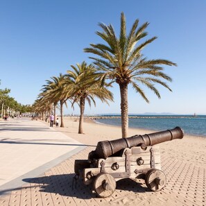 Un canon placé sur une promenade pavée, près de la plage, avec des palmiers et un ciel bleu dégagé.