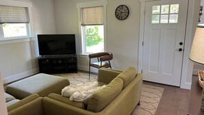 A living room with a sofa, ottoman, TV, and a door with a clock. - NEW Bungalow - Fenced yard - 30+ day rental - View of Lookout Mountain - Chattanooga (Chattanooga)
