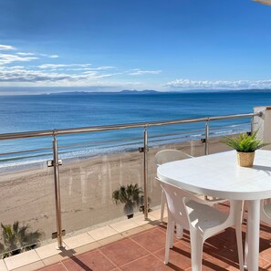 A balcony with a white table and chairs, overlooking a beach with a clear view of the sea and distant mountains. - Europa 62 in Roses With 2 Bedrooms and 2 Bathrooms (Roses)