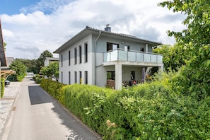 Une maison moderne à deux étages, dotée d’un balcon, d’une allée pavée et d’une végétation luxuriante.