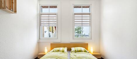 Un lit simple avec deux tables de chevet, une tête de lit en bois et deux fenêtres équipées de stores.