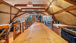 Un interior con una larga mesa de madera, zona de cocina con estufa y refrigerador, y vigas de madera.