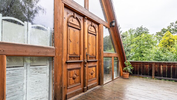 Una terraza de madera con un gran puerta de madera, una planta en maceta y vistas a árboles.