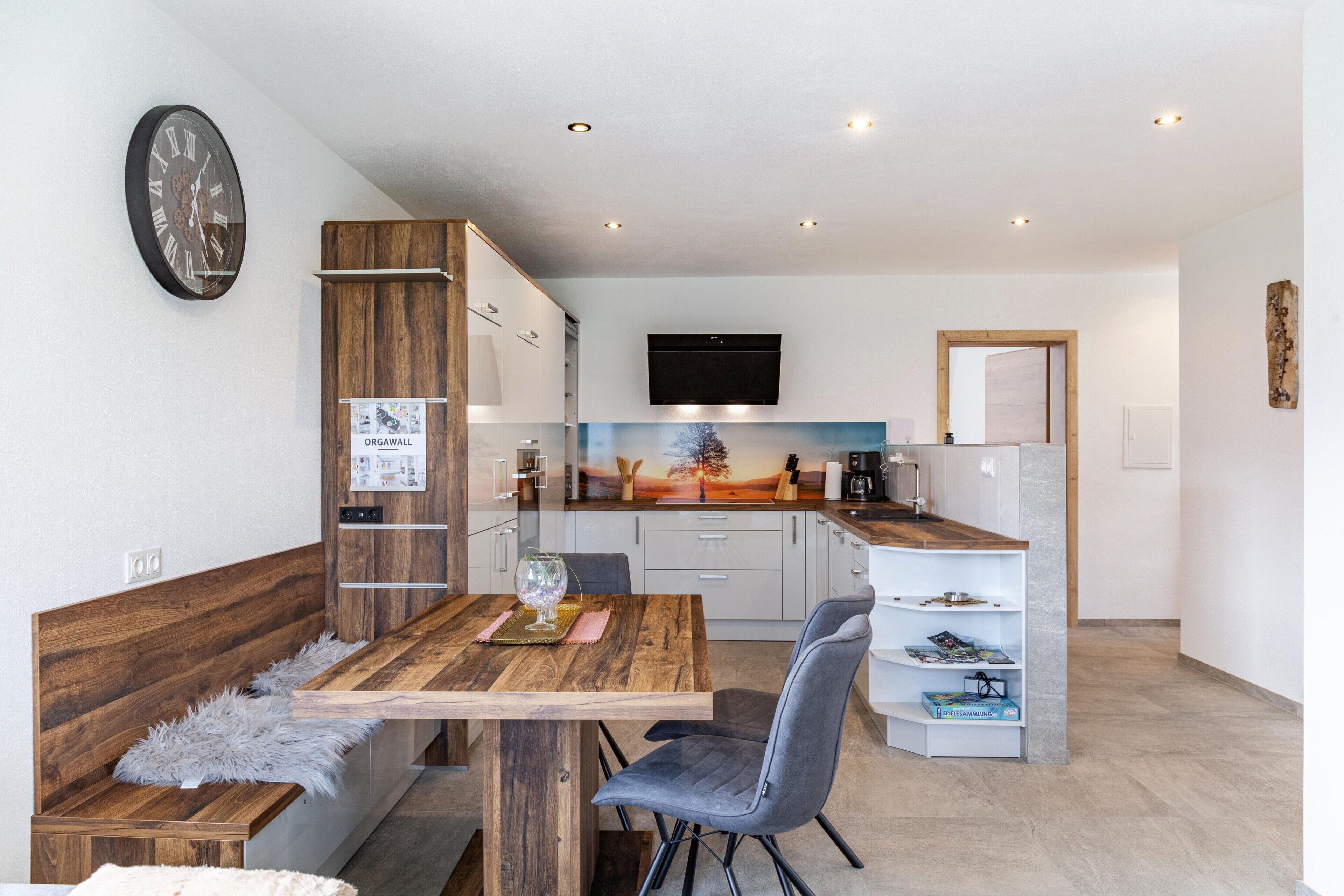 A modern kitchen with a wooden table, grey chairs, and a mounted flat-screen TV.