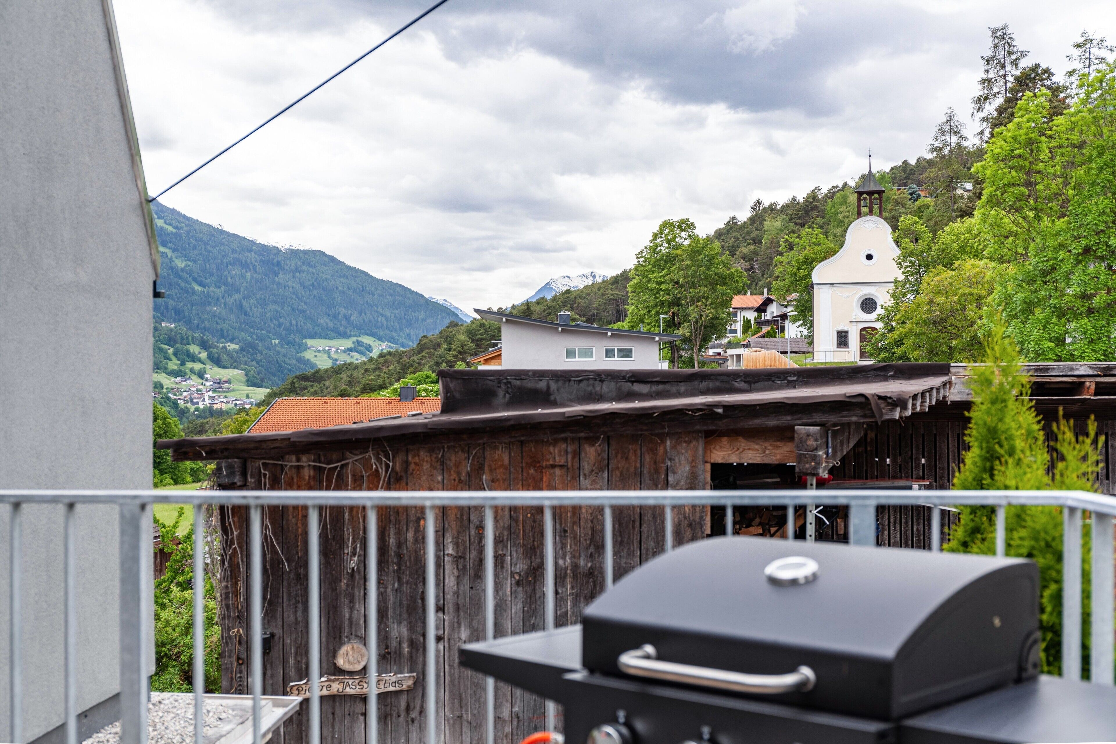 A balcony with a grill, a rustic wooden fence, and a view of a church and mountains in the background.
