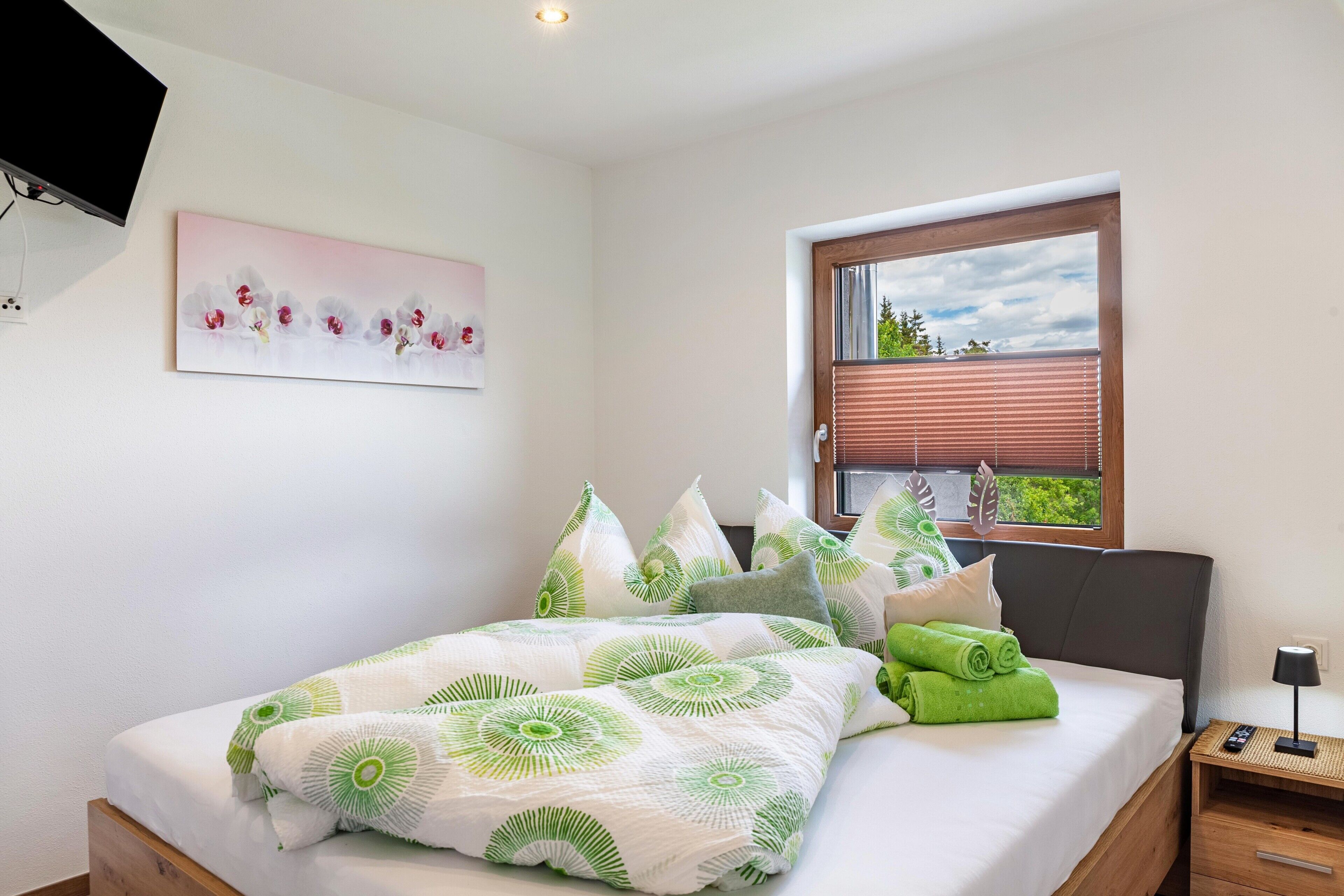 Una cama bien hecha con ropa de cama verde y blanca, un cuadro de flores en la pared y una ventana con persianas.