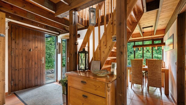 Ein Holzinterieur mit einer großen Tür, einer Holzkommode, Korbstühlen und Blick durch große Fenster.