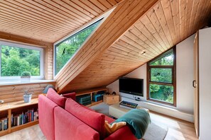 A modern loft bedroom with a sloped ceiling, large windows, a red sofa, a flat-screen TV, and a bookshelf. - Holiday Home 'Karlsminde' with Private Terrace, Garden, and Wi-Fi (Waabs)