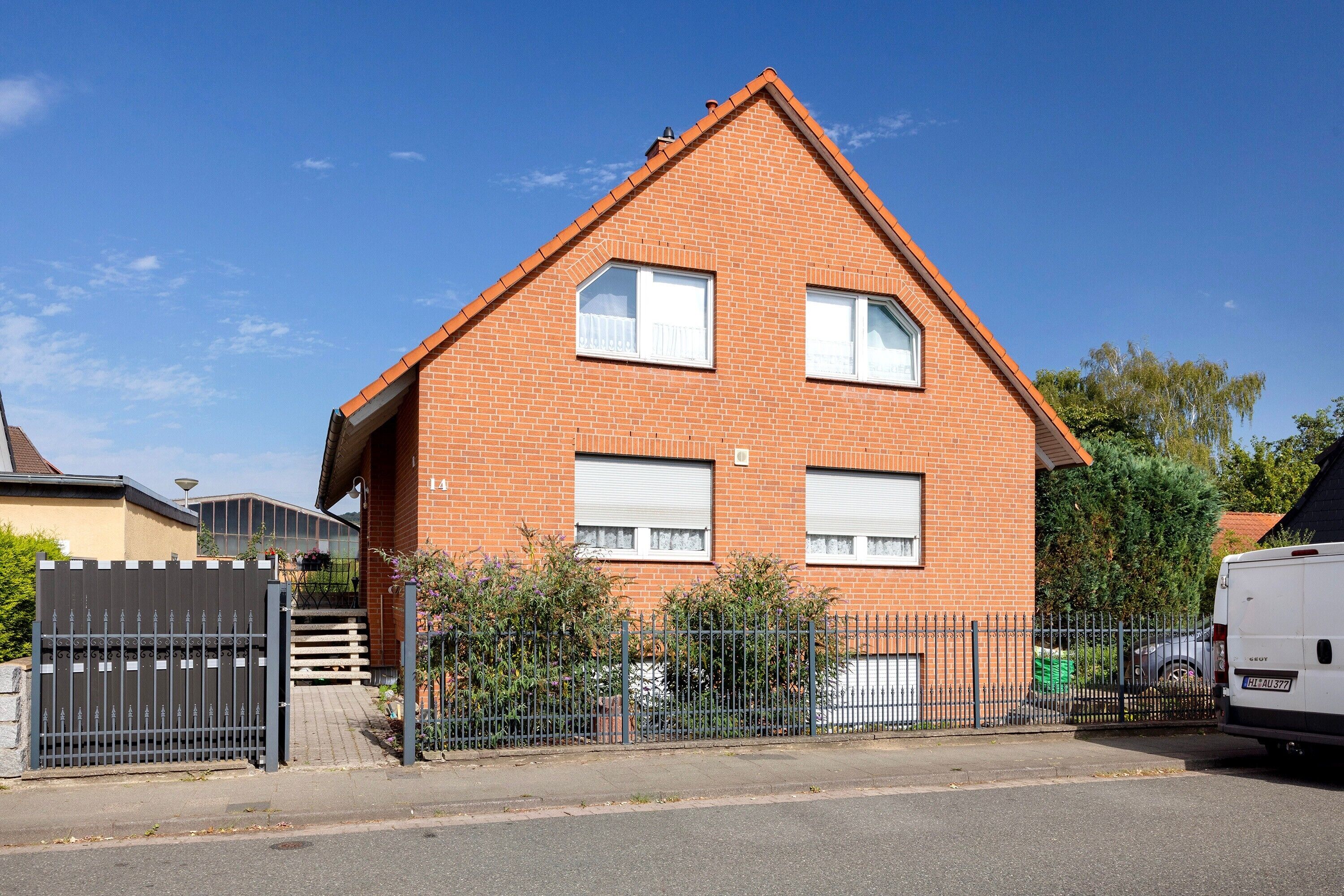 A two-story brick house with a gated driveway, a white van parked to the side, and a clear blue sky.