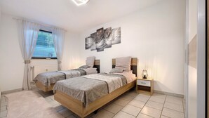Une chambre avec deux lits, une table de chevet, une commode, une fenêtre avec des rideaux et une œuvre d’art accrochée au mur.