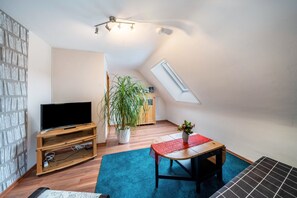A small, well-lit room with a television on a wooden stand, a potted plant, a dining table with a red table runner, and a slanted ceiling with a skylight. - Apartment 'Burgblick' with Mountain View, Shared Garden and Wi-Fi (Hausach)