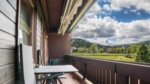 Un balcon en bois, agrémenté d’une table et de chaises, qui offre une vue sur un paysage verdoyant, avec des arbres et une colline.