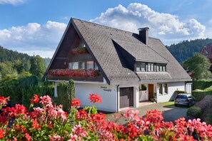 A house with a sloped roof, a balcony with flowers, a car parked in front, and a mountainous backdrop.