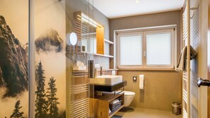 Een badkamer met een douchecabine van glas, een wastafel met spiegel, een toilet en een raam met zonwering.