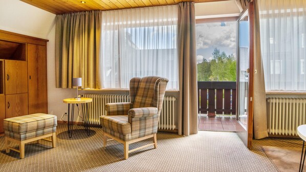 Een kamer met een gestreept fauteuil, een klein rond tafeltje met een lamp, een houten kast en uitzicht op het landschap door een groot raam.