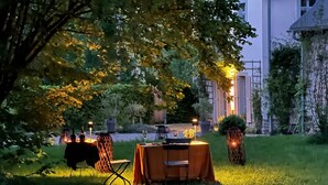Un área de comedor al aire libre con mesas y sillas, rodeada de vegetación y un edificio al fondo.
