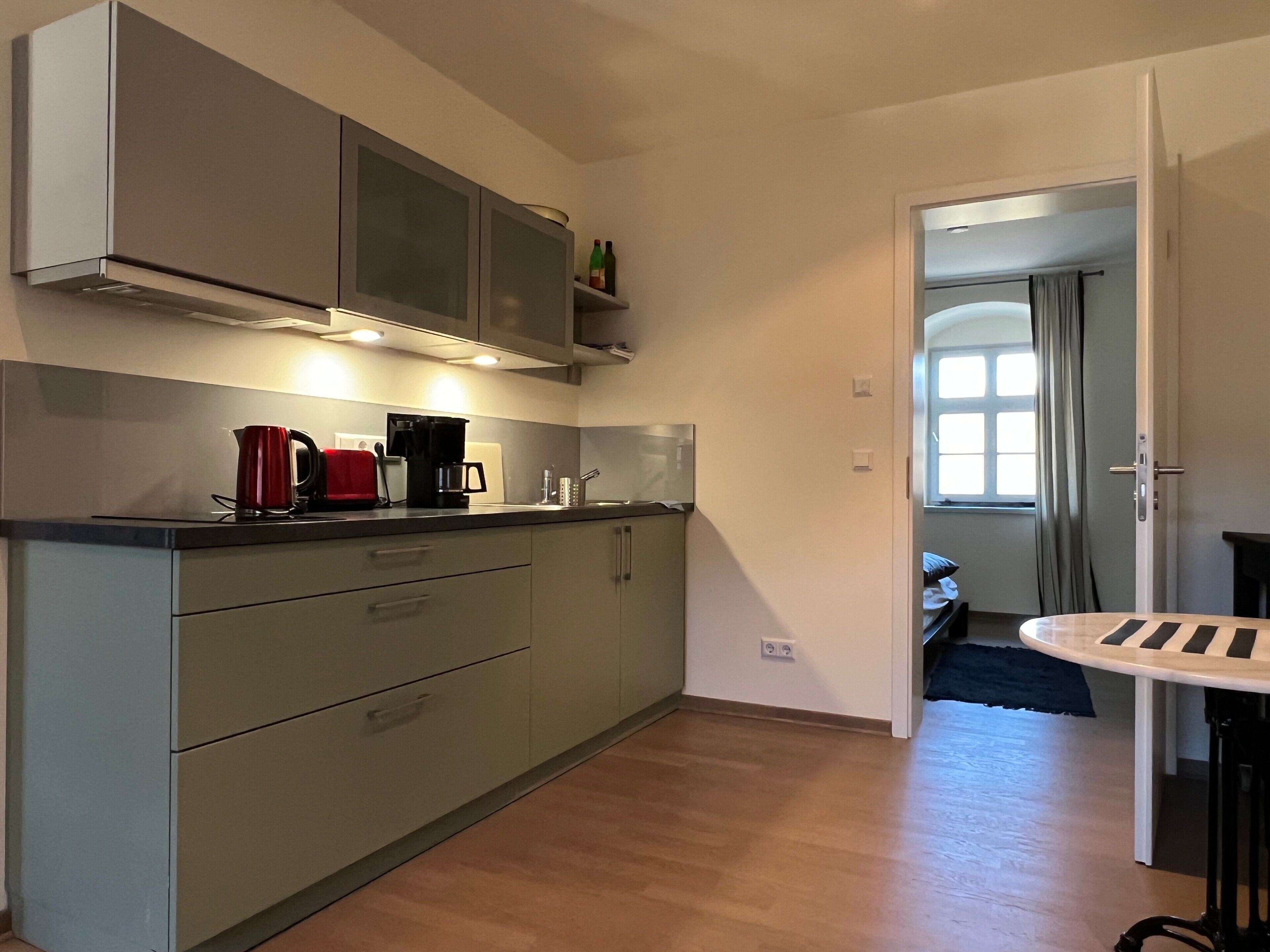 Eine moderne Küche mit grauen Schränken, einer Kaffeemaschine und einem Toaster. Ein Fenster mit Vorhängen und eine Tür zu einem anderen Raum.