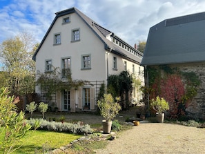 Un bâtiment à deux étages, doté d’un toit en pente, de nombreuses fenêtres et d’un jardin agrémenté de diverses plantes et d’un chemin de gravier.