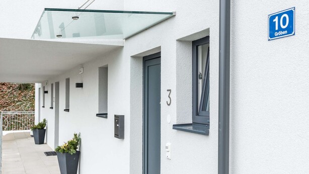 A modern building with a glass balcony, numbered doors, and potted plants.
