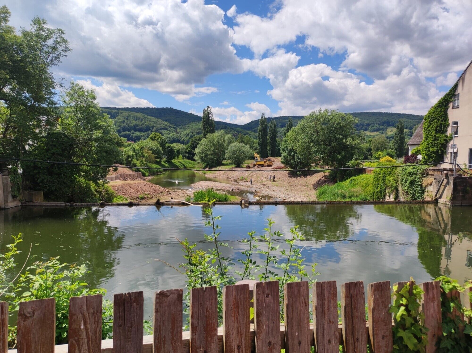 A river with a wooden fence in the foreground, a construction site on the opposite bank, and a building with ivy on the right.