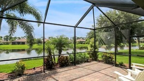 A covered patio with brick flooring and white chairs, overlooking a lake and greenery.