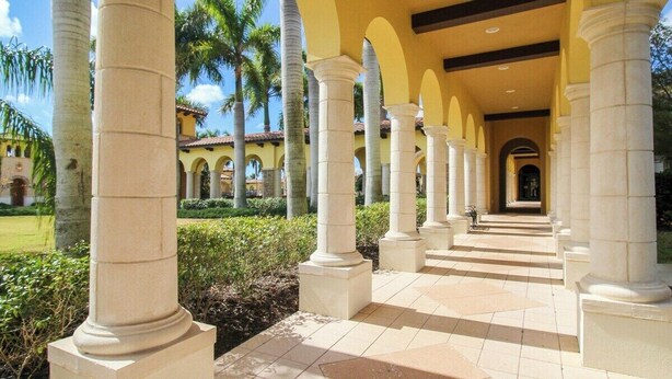 A covered walkway with columns, palm trees, and a building in the background.