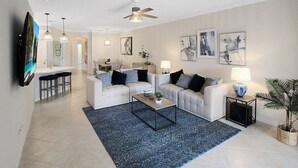 A modern living room with two sofas, a coffee table, and a TV. There are two pendant lights, a ceiling fan, and artwork on the walls.