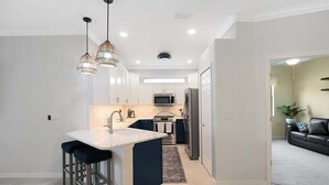 A modern kitchen with a central island, bar stools, pendant lighting, and a view into the living area.