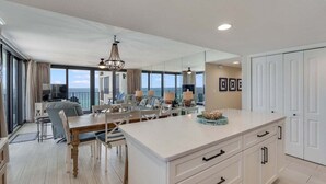 A spacious living area with a large dining table, a kitchen with white cabinetry, and a balcony with a view of the ocean.