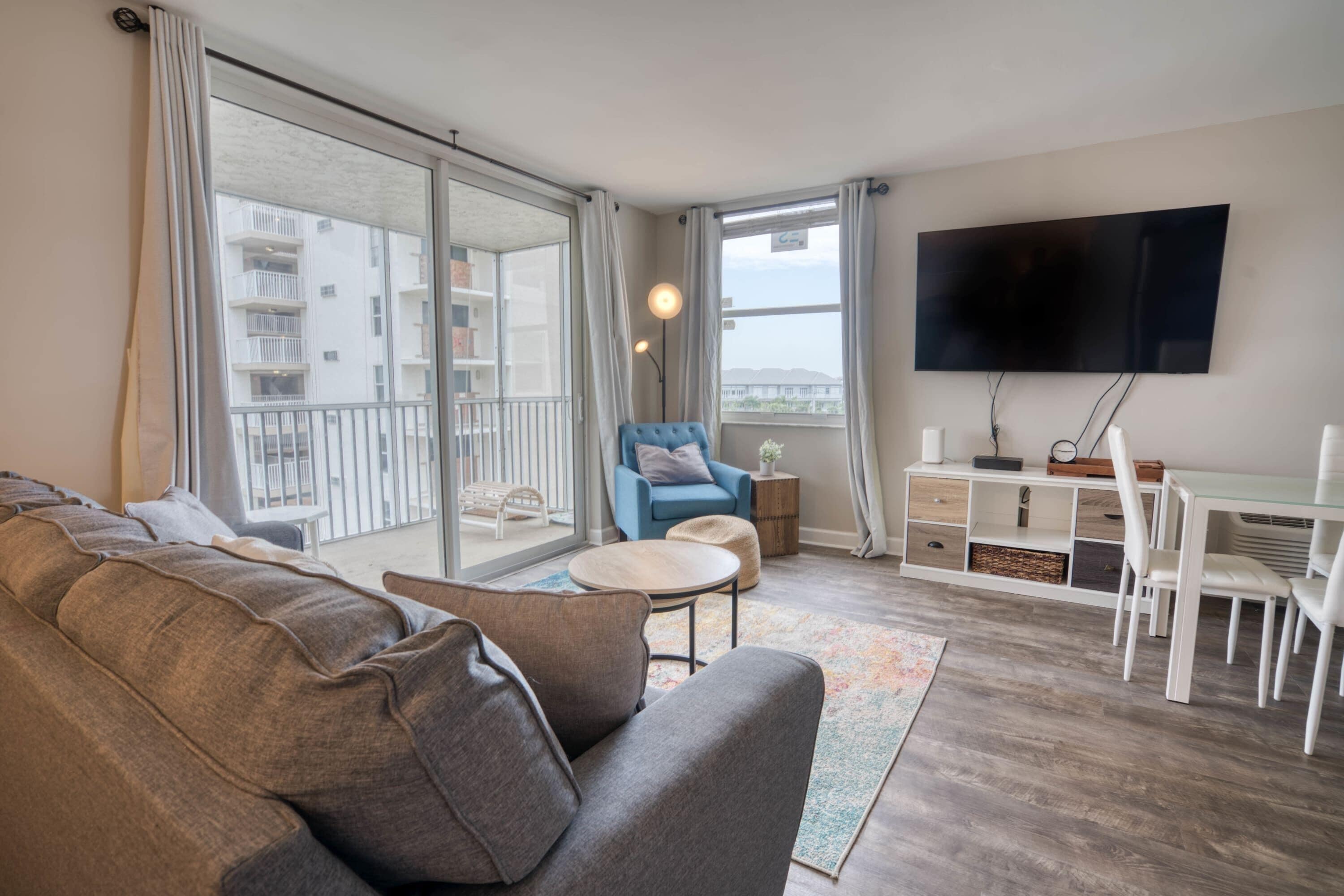 Ein modernes Wohnzimmer mit einer Couch, einem blauen Sessel, einem kleinen runden Tisch, einem an der Wand befestigten Fernseher und einem Balkon mit Blick auf die Stadt.