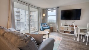 Un soggiorno moderno con un divano, una poltrona blu, un tavolino rotondo, una TV a parete e un balcone con vista sulla città.