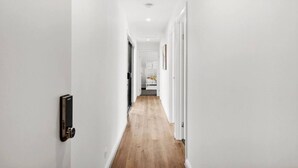 A narrow hallway with white walls and wooden flooring, leading to a room with a bed and a nightstand. - The Absolute Beach Escape For 21 (St Leonards)