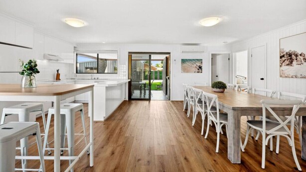 A modern kitchen with white cabinetry, a wooden dining table, and white bar stools. - The Absolute Beach Escape For 21 (St Leonards)