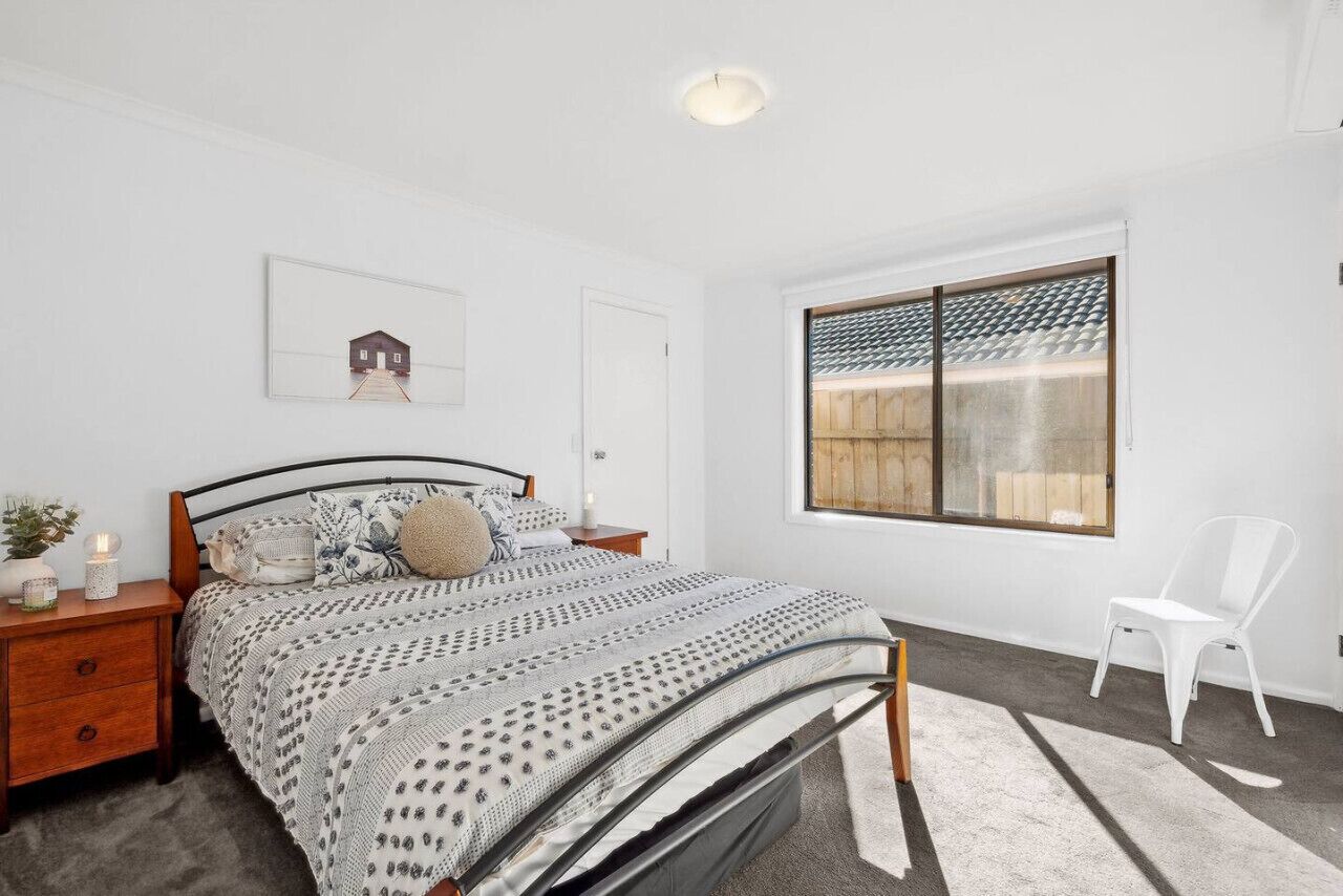 A bedroom with a bed, a nightstand, a chair, a window, and a framed picture on the wall.