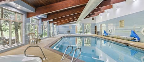 An indoor swimming pool with clear glass walls, wooden beams, and a view of trees outside.