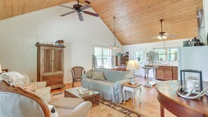 A spacious living room with a high ceiling, wooden floors, and a large window. There is a dining area with a table and chairs, and a kitchen in the background. - Charming Lawley Cottage: Deck, Fire Pit & Yard! (Lawley)