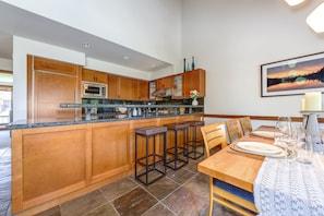A modern kitchen with wooden cabinets, a central island with bar stools, and a dining area with a table set for six. - Montebello 4864 - Private Hot Tub, Air Conditioning, Short Walk to the Village (Whistler)
