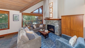 A spacious living room with a stone fireplace, wooden ceiling, and large windows offering a view of the outdoors. - Montebello 4864 - Air Conditioning, Private Hot Tub, Mountain Views, Close to Village (Whistler)