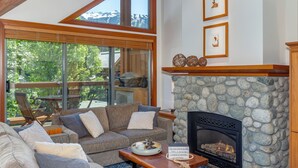 A cozy living room with a stone fireplace, a large window overlooking a deck, and a comfortable seating area. - Montebello 4864 - Air Conditioning, Private Hot Tub, Mountain Views, Close to Village (Whistler)