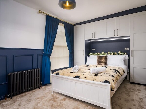 Ein Schlafzimmer mit einem Bett, weißen Schränken, einem Heizkörper und einem Fenster mit blauen Vorhängen.