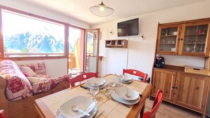 A dining area with a table set for four, a sofa with patterned cushions, a wooden cabinet with glass doors, and a television mounted on the wall. - Bright studio for 4 people with magnificent view (Les Deux Alpes)