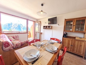 A dining area with a table set for four, a sofa with patterned cushions, a wooden cabinet with glass doors, and a television mounted on the wall.
