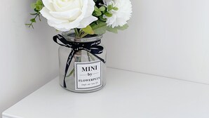 A white shelf with a glass jar containing white flowers and greenery, labeled "MINI by FLOWERPLUS." - 3rd Alley Inn (Kaohsiung)