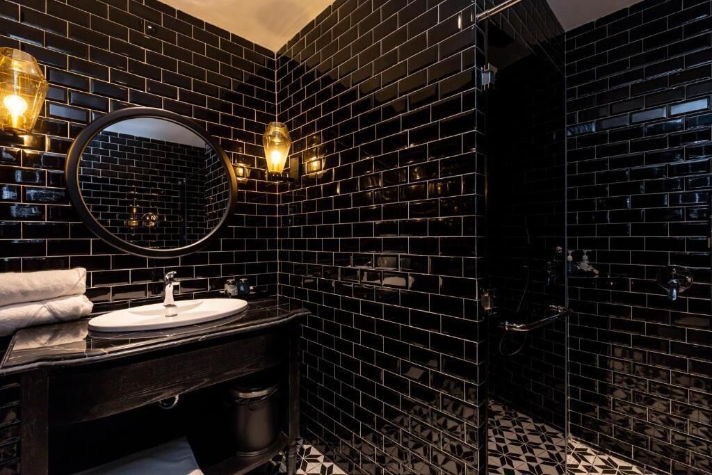 Ein modernes Badezimmer mit schwarzen Fliesenwänden, einem runden Spiegel, einem Waschbecken und einer Duschkabine.
