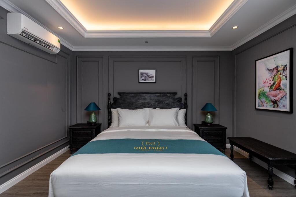 Een hotelkamer met een groot bed, twee nachtkastjes met lampen, een bankje, een wandairconditioner en een inlijsting foto aan de muur.
