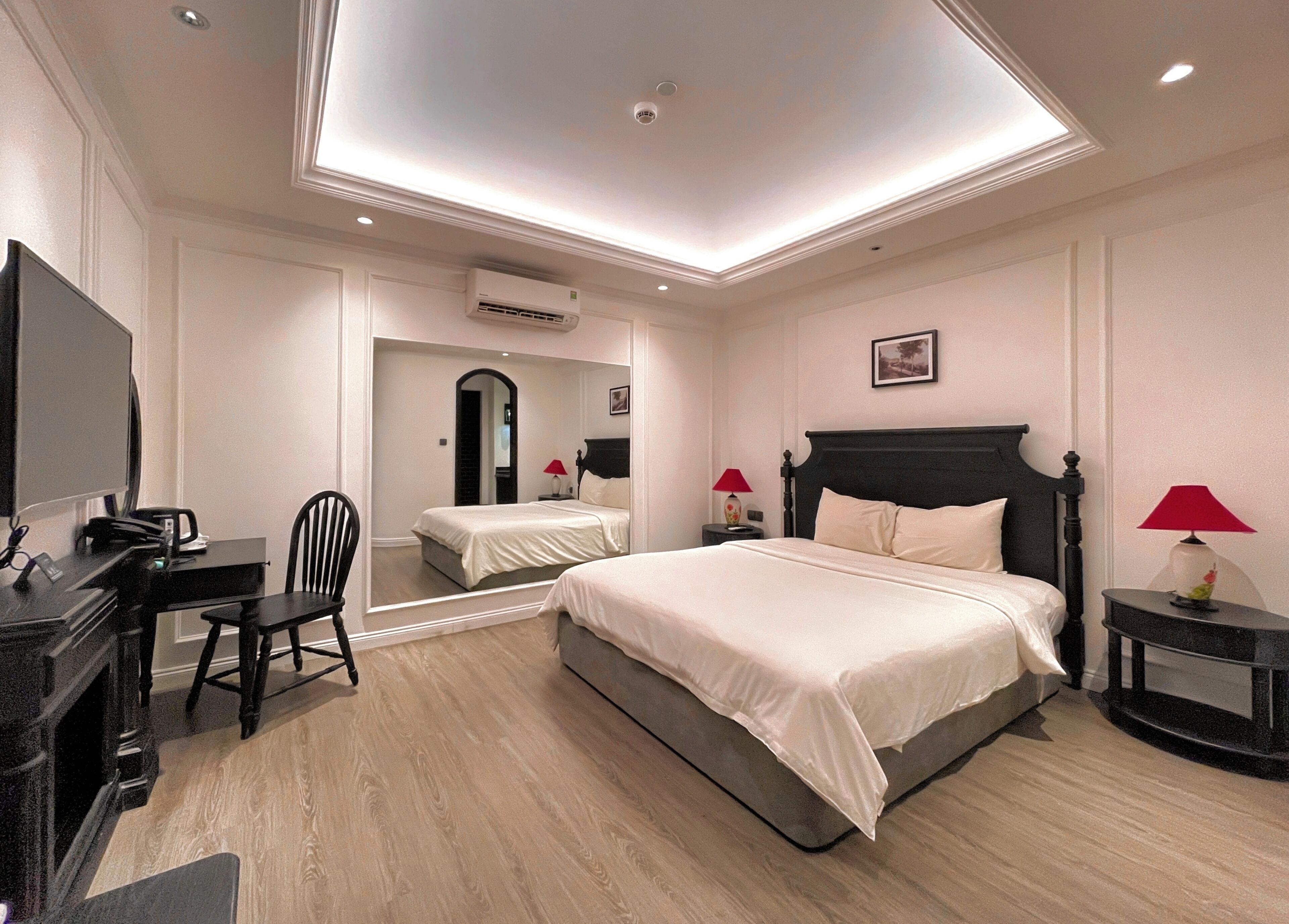 A hotel room with a bed, a desk with a chair, a TV, a mirror, and a ceiling light.