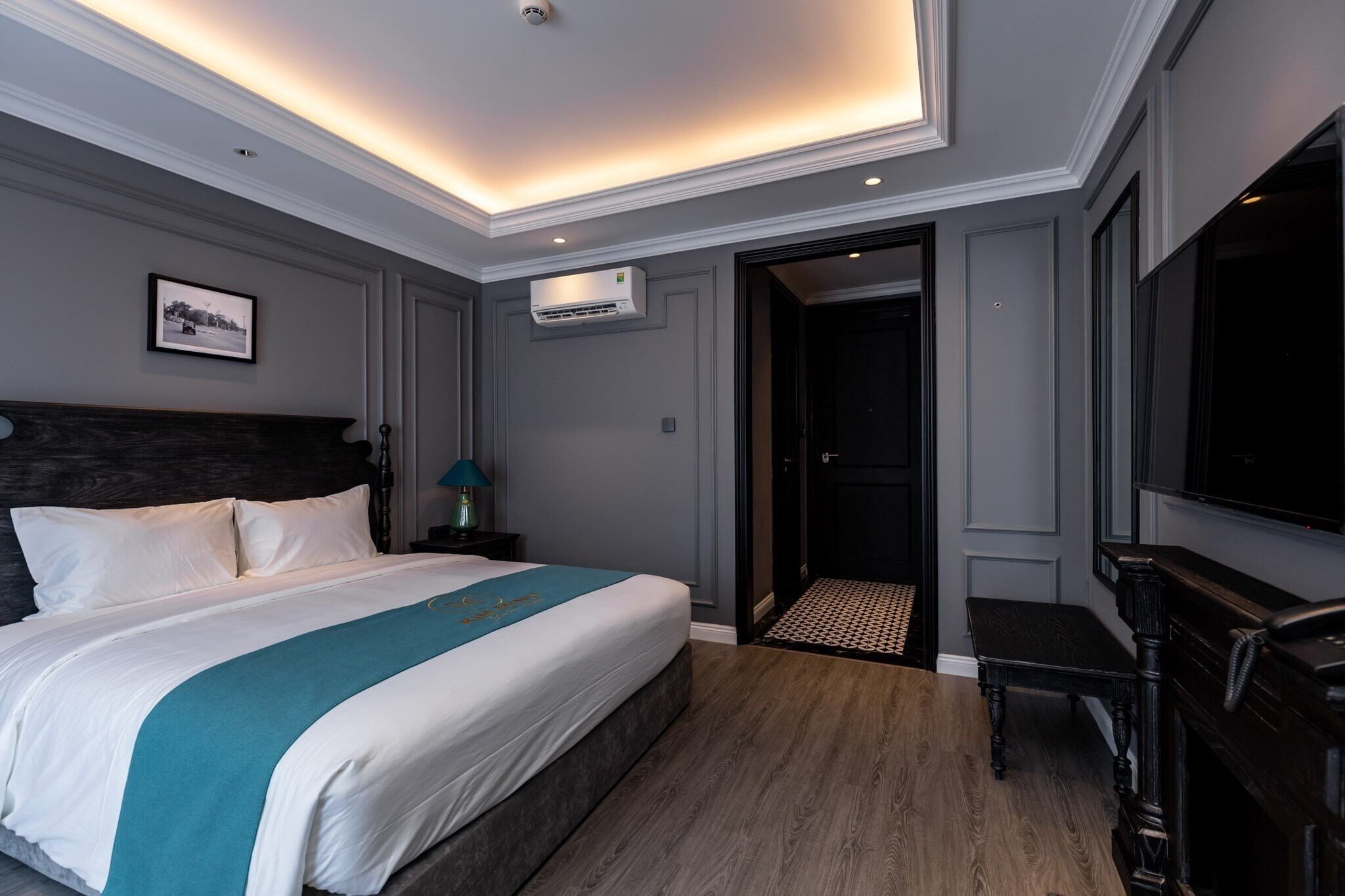 A hotel room with a large bed, a bedside table with a lamp, a wall-mounted air conditioner, and a door leading to another room.