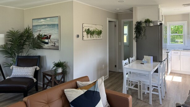 A modern living room with a brown leather sofa, a white dining table with chairs, and a kitchen area with white cabinets and a window. - The Oar Cottage (24) · walk Mystic-Train/EV Lvl-2 (Stonington)