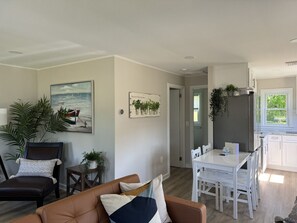 A modern living room with a brown leather sofa, a white dining table with chairs, and a kitchen area with white cabinets and a window. - The Oar Cottage (24) · walk Mystic-Train/EV Lvl-2 (Stonington)