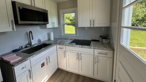 A kitchen with white cabinets, a stainless steel microwave, a sink, and a window providing a view of the outdoors. - The Oar Cottage (24) · walk Mystic-Train/EV Lvl-2 (Stonington)